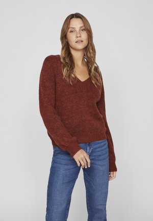 Rust-colored v-neck sweater in a soft, textured knit. Long sleeves, relaxed fit, paired with blue denim jeans. Simple design, no patterns.