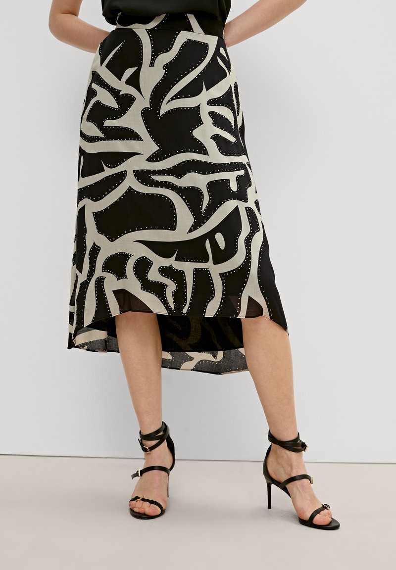 Zwart-wit gemusterde midi-rok met een high-low zoom, met abstracte ontwerpen en veiligheidsspelden als accenten. Draagt met zwarte sandalen met straps.