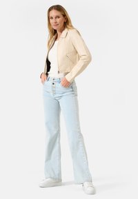 Beige leren jas met een kraag, ritssluiting en zijzakken. Lichtblauwe high-waisted flare jeans met knopen aan de voorkant, gecombineerd met witte sneakers.