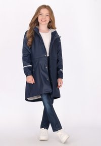 Jūrinis vandeniui atsparus lietpaltis su pilku gobtuvu, spaudžių mygtukų ir atspindinčių akcentų, derinamas su mėlynais džinsais ir baltais sportbačiais.