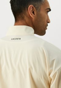 Man in beige Lacoste polo shirt shown from the back and side, focusing on the shirt's upper back and collar label.