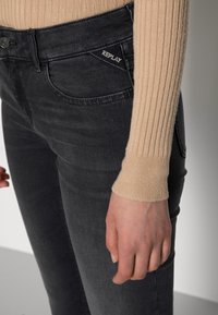 Svarta denimjeans med slim fit och subtil struktur, med en REPLAY-logga broderad på framfickan. Ljusbeige ribbad ärm synlig.
