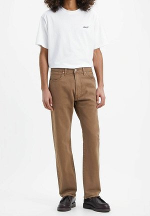 Personne portant un t-shirt blanc Levi's, un jean marron droit et des mocassins en cuir noir, debout sur un fond blanc.