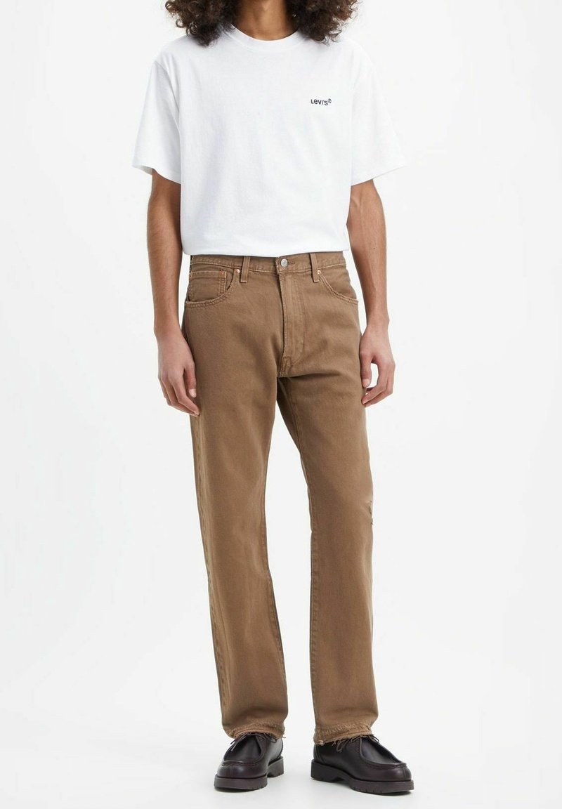Personne portant un t-shirt blanc Levi's, un jean marron droit et des mocassins en cuir noir, debout sur un fond blanc.