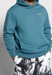 Sudadera azul de Calvin Klein llevada sobre pantalones con un patrón blanco y negro, con las manos en los bolsillos.