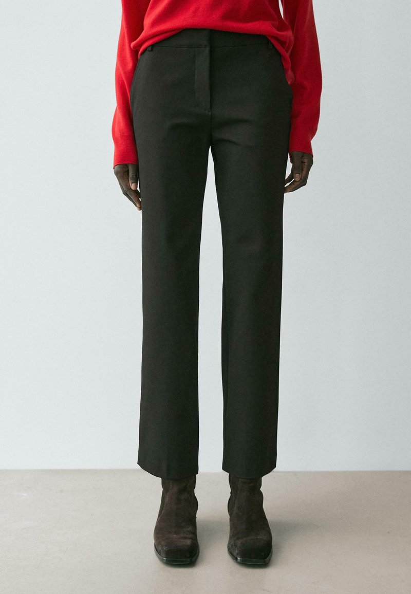 Schwarze, maßgeschneiderte Hose mit geradem Schnitt, glatter Textur, vorderen Bundfalten und verdecktem Bundabschluss, kombiniert mit braunen Stiefeln.