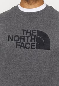 Grauer Sweatshirt mit Rundhalsausschnitt, auf der Brust mit einem großen schwarzen, gestickten "THE NORTH FACE" Logo. Weiche Textur.