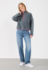 Gray fleece pullover with a high collar and pink zipper. Features an open kangaroo pocket and contrasting pink hem. Worn with blue jeans.