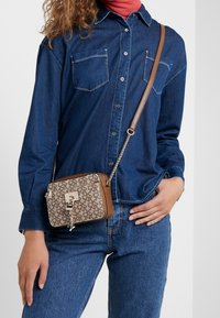 Hnědo-beige crossbody taška s geometrickým vzorem, zlatými detaily, zipovým zapínáním a řetízkem, držena proti denimové košili.