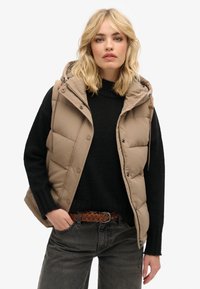 Beige Daunenweste mit Kapuze, ausgestattet mit einem Reißverschluss und Druckknöpfen. Über einem schwarzen Strickpullover gestylt, kombiniert mit dunklen Jeans und einem braunen Gürtel.