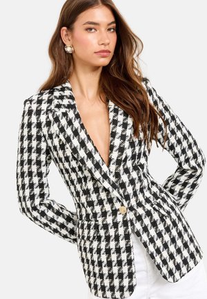 Mladá žena nosí čierno-biely sako károvaný vzor houndstooth s jediným zlatým gombíkom, v kombinácii s bielymi nohavicami a veľkými okrúhlymi naušnicami.