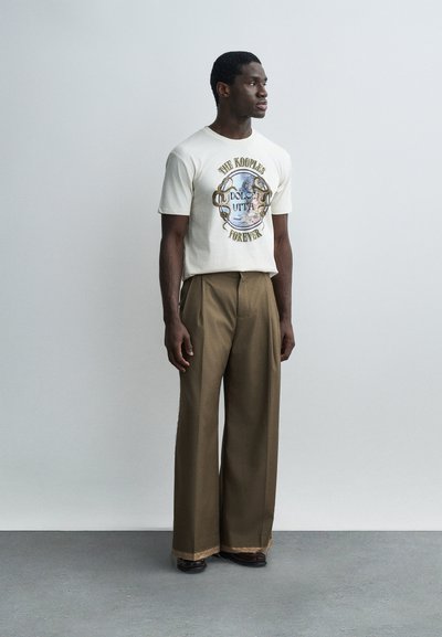 Άνδρας στέκεται κοιτώντας δεξιά, φορώντας λευκό γραφικό μπλουζάκι με κείμενο και καφέ παντελόνι με φαρδιές μπατζάκια, μπροστά σε απλό φωτεινό φόντο.