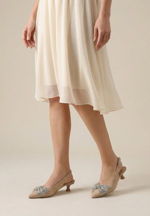 Vrouw die beige slingback hakken met strikjes van rhinestones draagt en een vloeiende crème midi-rok, tegen een neutrale achtergrond.