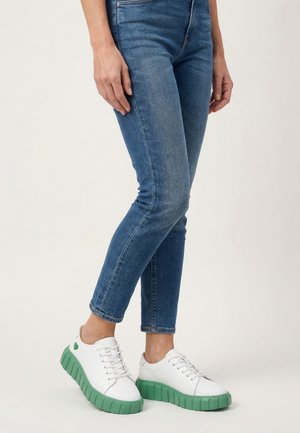 Person wearing blue skinny jeans and white sneakers with thick green soles, standing against a plain light background.