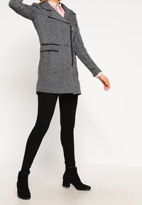 Abrigo gris de espiga con cuello con muescas, cremallera asimétrica y bolsillos frontales. Combinado con pantalones pitillo negros y botas de tobillo.