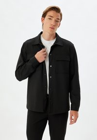 Camicia nera con bottoni, due tasche sul petto, maniche lunghe e taglio dritto. Indossata sopra una maglietta bianca. Tessuto morbido al tatto.
