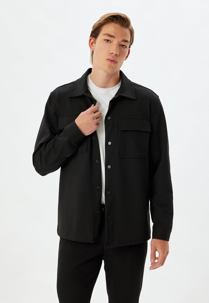 Camicia nera con bottoni, due tasche sul petto, maniche lunghe e taglio dritto. Indossata sopra una maglietta bianca. Tessuto morbido al tatto.