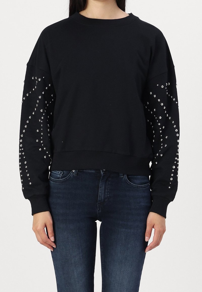Femme portant un sweat-shirt noir à manches longues décoré de clous argentés sur les deux manches, associé à un jean bleu foncé.