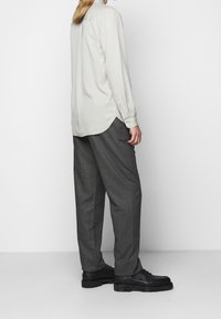 Camicia beige chiaro a maniche lunghe con orlo arrotondato e polsini con bottone, abbinata a pantaloni check grigio scuro e scarpe nere in tessuto.