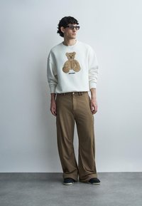 Jeune homme avec des lunettes de soleil portant un sweat-shirt blanc avec un motif d’ours en peluche et un pantalon large marron, debout contre un mur uni.