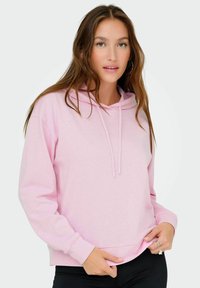 Hanorac roz deschis cu sireturi, mâneci lungi și o croială lejeră. Material moale cu manșete și tiv cu dungi. Parte ascunsă în pantaloni negri.