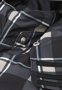Sort og grå plaid-stof med sølvtrykknapper og sorte accentremme. Materialet fremstår glat med en let glans.