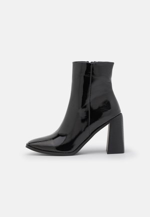 Bottine en cuir verni noir avec bout pointu, fermeture éclair latérale et talon bloc haut et épais sur un fond clair uni.