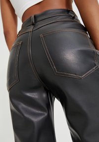 Pantalons taille haute en simili cuir noir avec des coutures brunes contrastantes et deux poches arrière, présentant une texture lisse et brillante.
