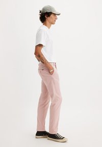 Pantaloni di cotone rosa chiaro con design a gamba dritta, tasche frontali e tasca posteriore a toppa. Abbinati a una t-shirt bianca e sneakers nere.