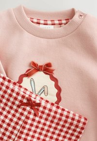 Conjunto de bebé rosa con cuello acanalado, lazos de terciopelo rojo, ilustración de orejas de conejo y pantalones a cuadros rojos y blancos con un lazo.