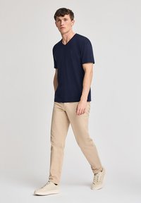 Granatowa koszulka V-neck, krótki rękaw, wykonana z miękkiej tkaniny. Połączona z beżowymi spodniami i jasnobeżowymi trampkami, stojąc na neutralnym tle.