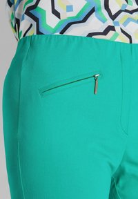 Grüne Hosen mit schmaler Passform, glatter Textur und einer Reißverschlusstasche auf der rechten Seite. Das Oberteil ist mit geometrischen Formen gemustert.