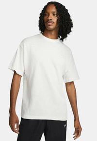 Vit bomull t-shirt med korta ärmar, rund halsringning och en liten blå logotyp på bröstet. Mjuk, avslappnad design, kombinerad med svarta byxor.