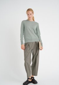 Maglione verde lavorato a maglia con scollatura rotonda, abbinato a pantaloni grigi a quadri e sandali neri con cinturini. Clutch tenuta nella mano destra.