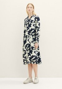 Femme portant une robe noire midi avec de grandes fleurs blanches imprimées et des chaussures à lacets de couleur crème, se tenant contre un fond clair et uni.