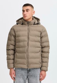 Beige polstret jakke med hætte, med en frontlynlås, elastiske snore og en kviltet tekstur. Båret over grå denimbukser.