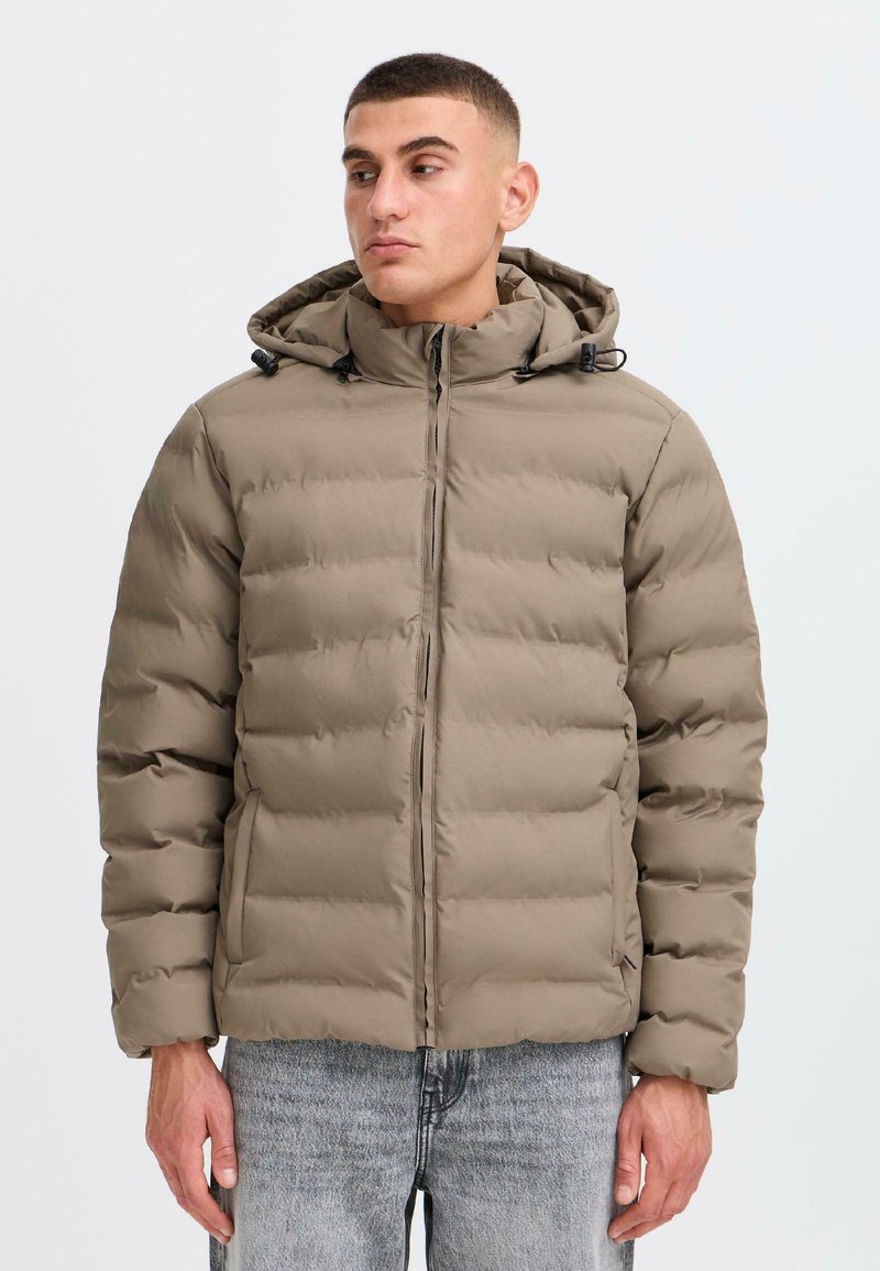 Beige polstret jakke med hætte, med en frontlynlås, elastiske snore og en kviltet tekstur. Båret over grå denimbukser.