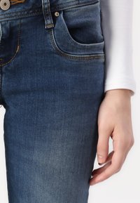 Dark blue denim jeans with a slightly faded texture, featuring a front pocket, orange stitching, and a silver button closure.