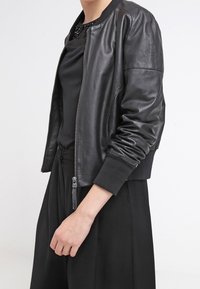 Giacca bomber in pelle nera con chiusura zip, polsini a coste e texture liscia. Indossata sopra un top scuro e pantaloni.