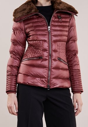 Frau trägt eine glänzende, kastanienbraune gesteppte Daunenjacke mit braunem Pelzkragen und schwarzen Hosen, vorderer Reißverschluss geschlossen, steht mit entspannten Händen.