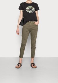 Svart T-shirt med ett flerfärgat läpptryck, olivgröna cargo-byxor med sidofickor och svarta flip-flops, hålls tillsammans med en liten svart väska.