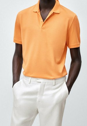 Man wearing a short-sleeve orange polo shirt tucked into high-waisted white pants, hands in pockets, standing against a plain background.