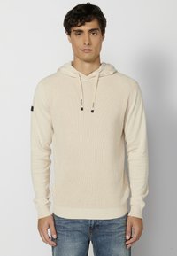 Haut à capuche tricoté de couleur crème avec un motif texturé, cordon de serrage ajustable et poignets côtelés. Associe avec un jean en denim bleu.