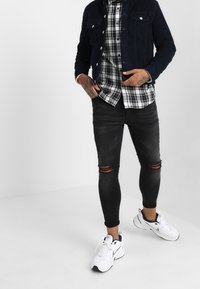 Pantalon skinny noir déchiré, chemise à carreaux avec des boutons, et veste en denim bleu marine. Des chaussures de sport blanches complètent l'ensemble.