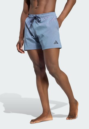 Męski model w jasnoniebieskich kąpielówkach Adidas ze sznurkiem i logo, stojący boso na jednolitym jasnym tle.