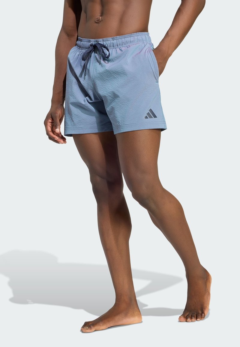 Modèle masculin portant un short de bain Adidas bleu clair avec cordon et logo, debout pieds nus devant un fond clair uni.