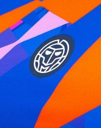 Tela azul y naranja con un patrón de formas geométricas. Presenta un logo negro circular con un diseño de cara estilizada en el centro.