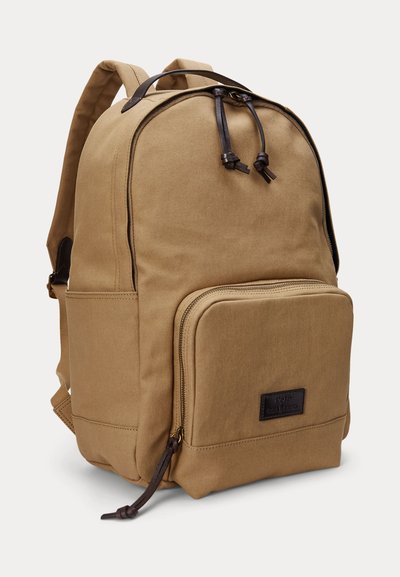 Béžový plátenný batoh s predným zipsovým vreckom, koženými ťahadlami zipsu, vystlanými ramennými popruhmi a horným nosným rukoväťou na bielom pozadí.