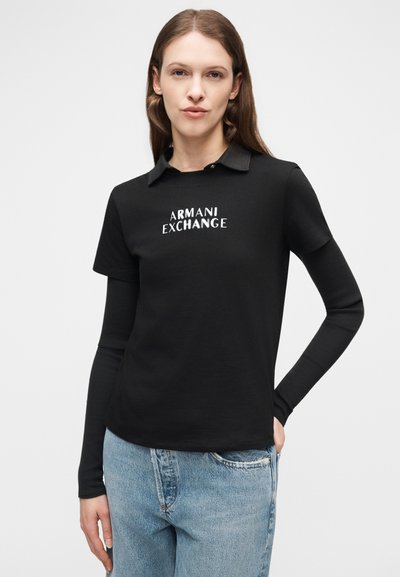 Γυναίκα που φοράει μαύρη μπλούζα με στρώσεις, μακριά και κοντά μανίκια, με το κείμενο «ARMANI EXCHANGE», συνδυασμένη με ανοιχτόχρωμο τζιν παντελόνι.