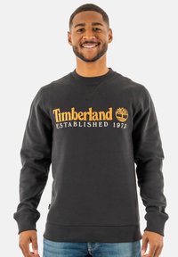 Zwarte sweatshirt van zacht materiaal met "Timberland Established 1973" in gouden borduursel en een boomlogo. Geribde mouwen en onderrand.